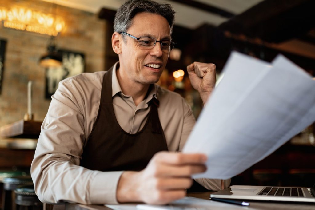  Cafe owner smiles as he sits at table and reviews small-business Cyber Insurance policy, laptop computer open on table. 