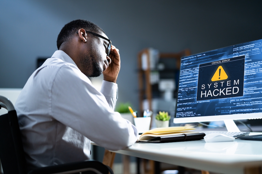 Businessman closes eyes and holds hand to head in despair as “SYSTEM HACKED” alert flashes on his computer monitor.