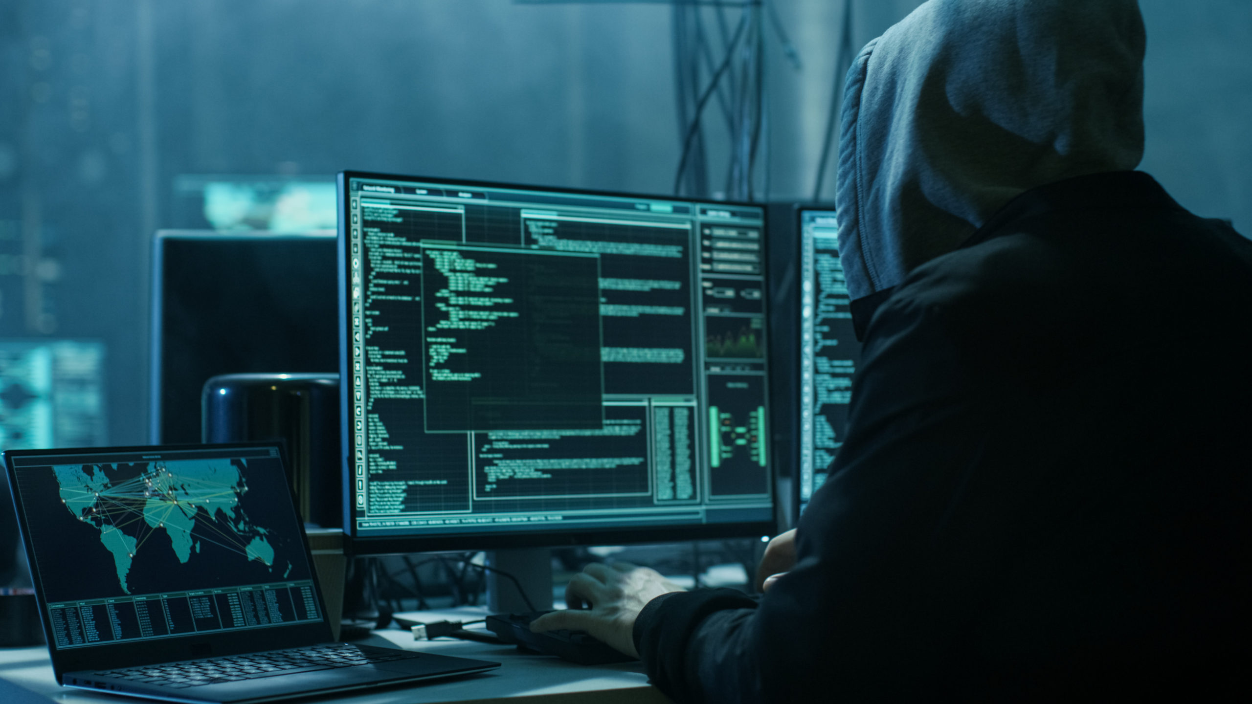 Hooded hacker in a dim, atmospheric warehouse. in front of multiple monitors and laptops attempting a data breach.