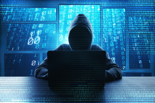 A cybercriminal in a dark hoodie sits in front of a laptop with blue code overlaying the image. 