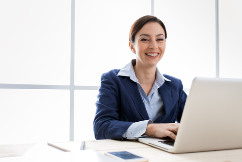 Smiling young business woman with brown hair and blue suit jacket sits at laptop computer.