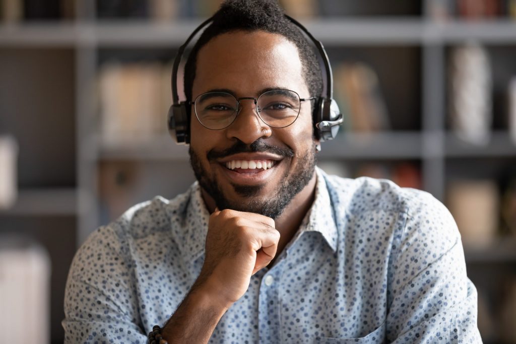 A smiling African American male leans on his hand while he takes a phone call from his headset at his desk. 