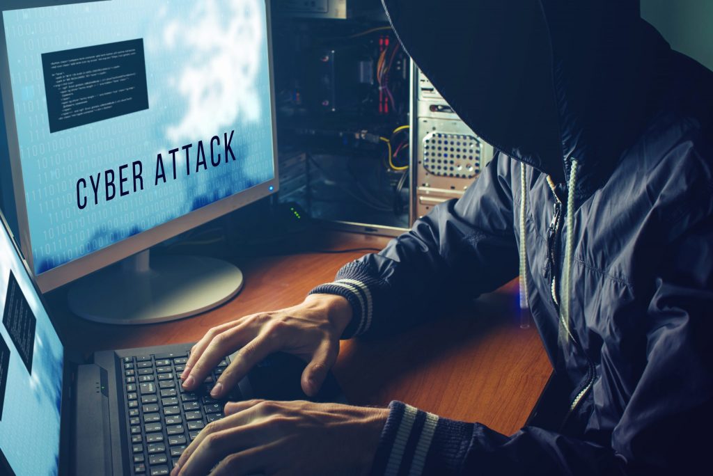 Hacker in black hoodie launches a cyber attack against a business from a dark room on a blue screen with code. 