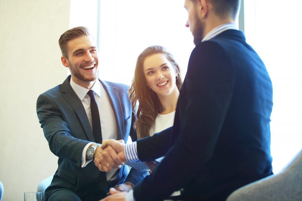 Three business professionals (two male and one female) smile and shake hands in a conference room.