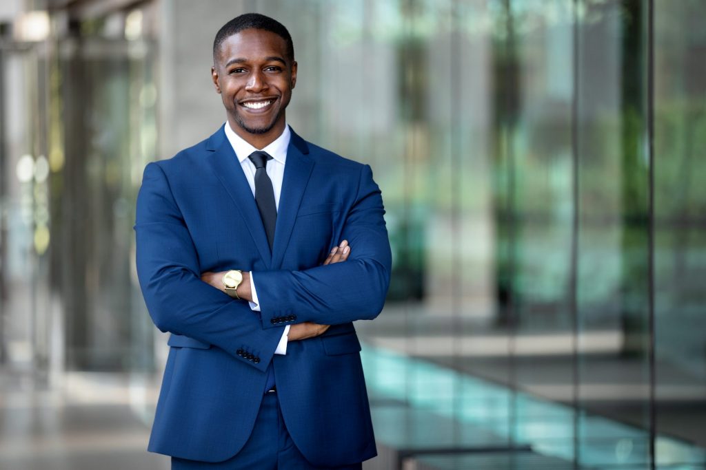 A young African American businessman wears a navy suit and smiles with his arms crossed. 
