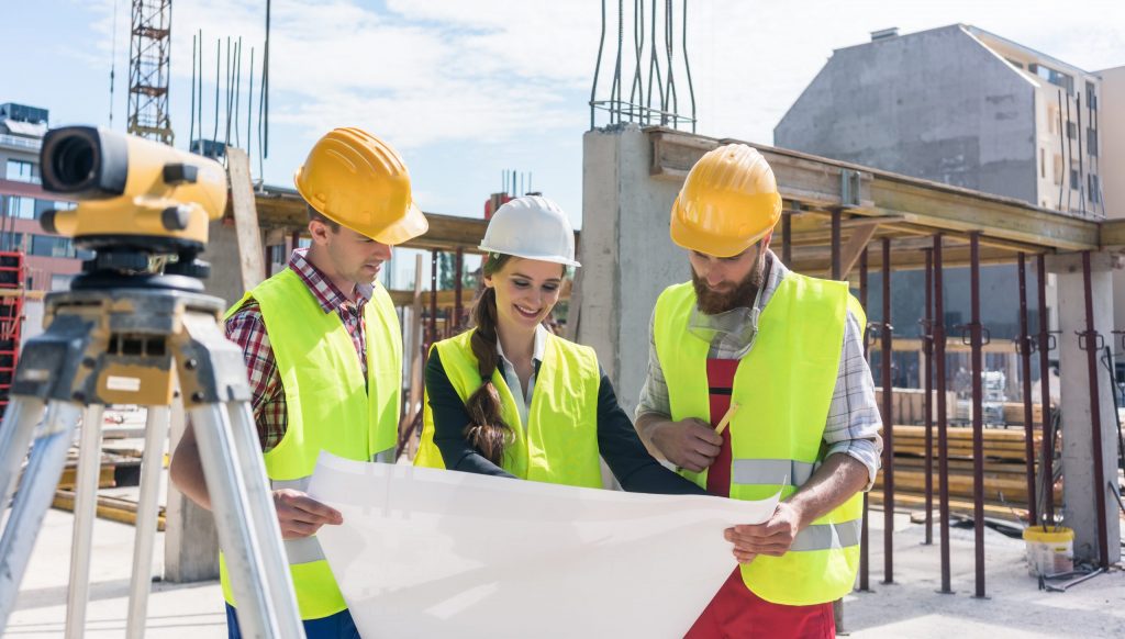 Female architect holds and discusses blueprint with two male construction managers at site, all wearing hard hats.