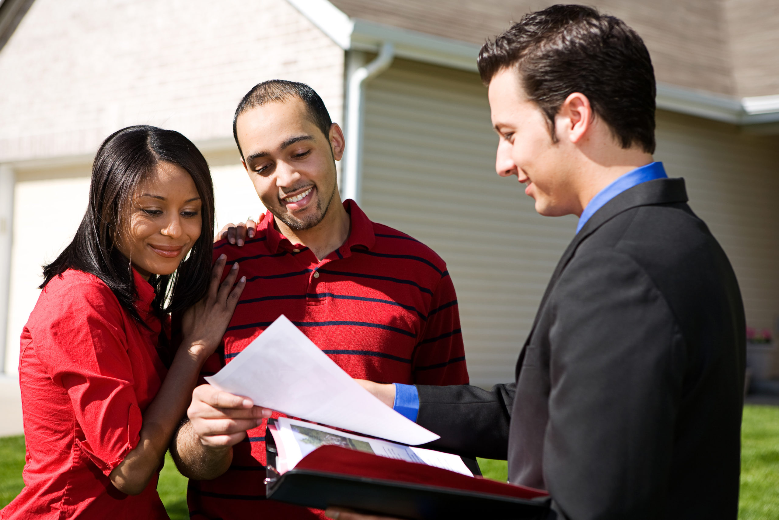 Male real estate agent stands outside newly sold house with man and woman, handing them completed home ownership paperwork.