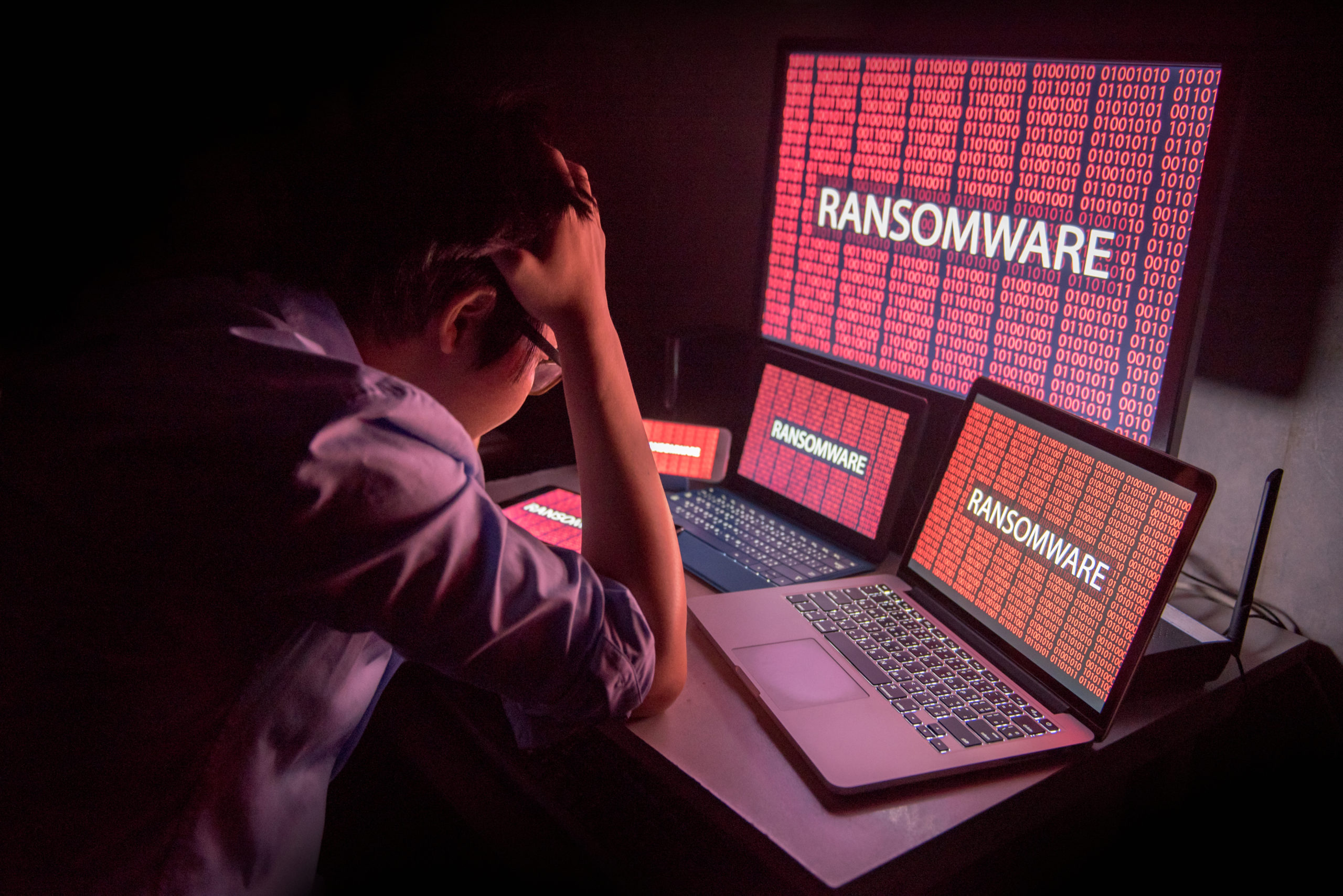Upset businessman sits in front of several laptop and tablet computers and large monitor, all displaying ransomware lock screens.