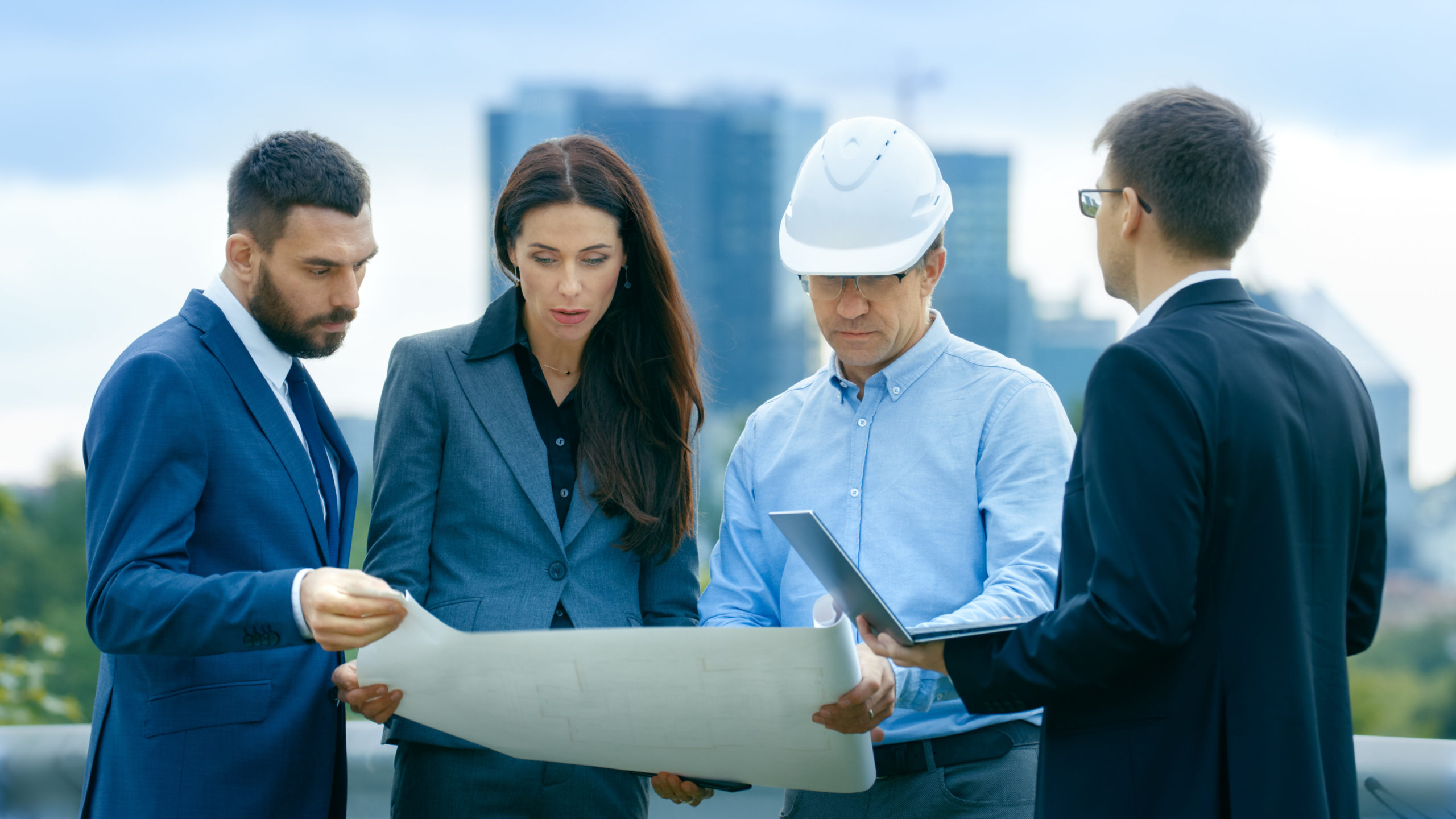 Real estate developer, general contractor, property manager, and investor holding laptop computer review blueprints at site.