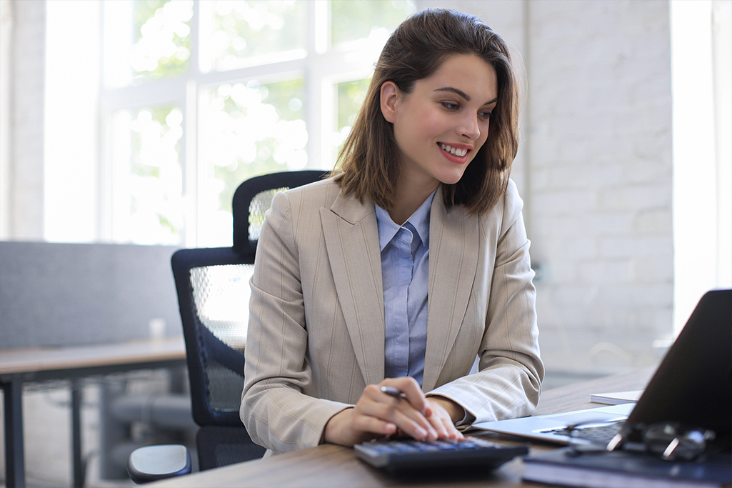 Retail broker sits at laptop computer to review insurance products for her clients, pen in hand, touching desktop calculator. 