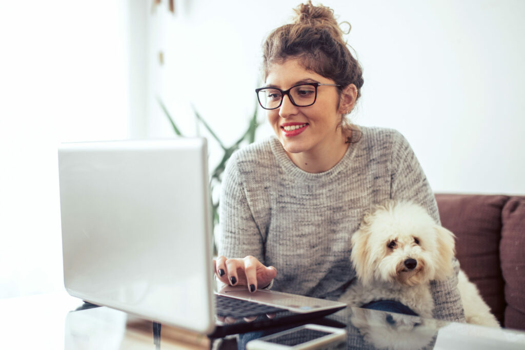 A young woman with her white dog on her lap works on her computer at home.
