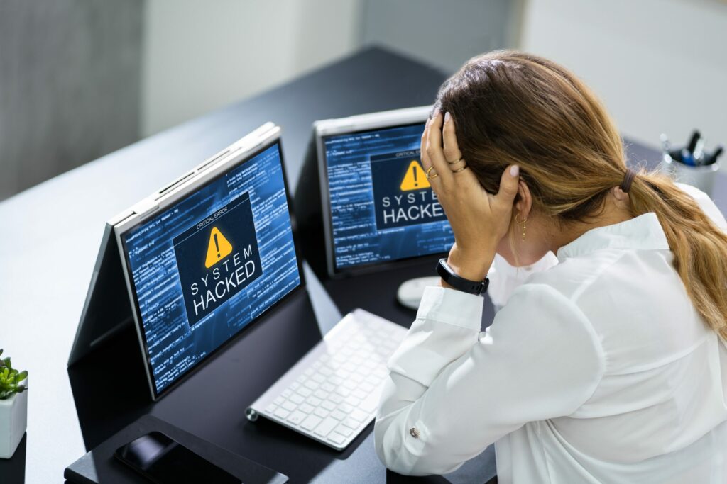 Businesswoman sits at desk, holding hands to her head, as “SYSTEM HACKED” messages flash on her two laptop monitors. 