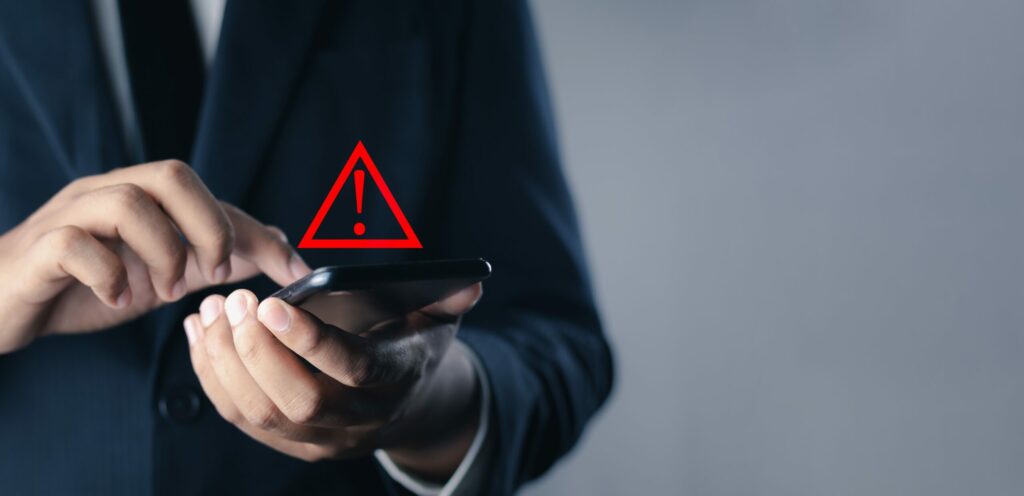 Businessman in suit holds smartphone; exclamation point in triangular graphic indicates his device has been hacked.